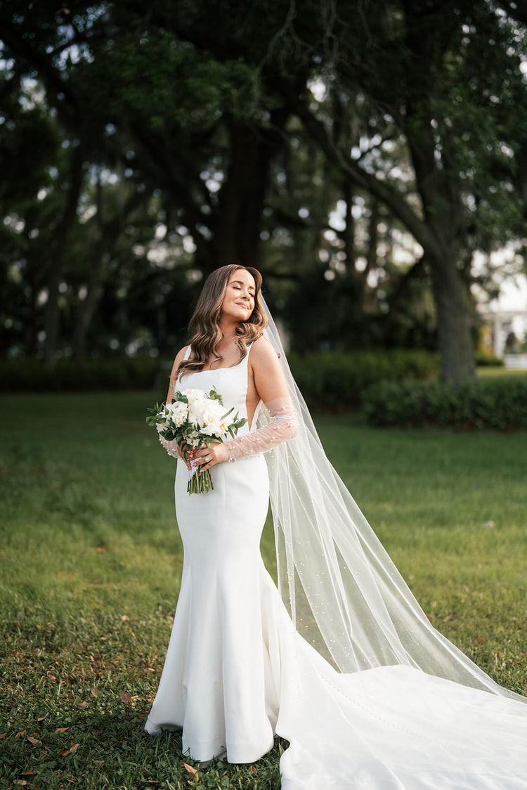 Bride stands alone on a lawn, eyes closed, pearl veil spread wide behind her, holding a white bouquet beneath tall oak trees, wedding photography
