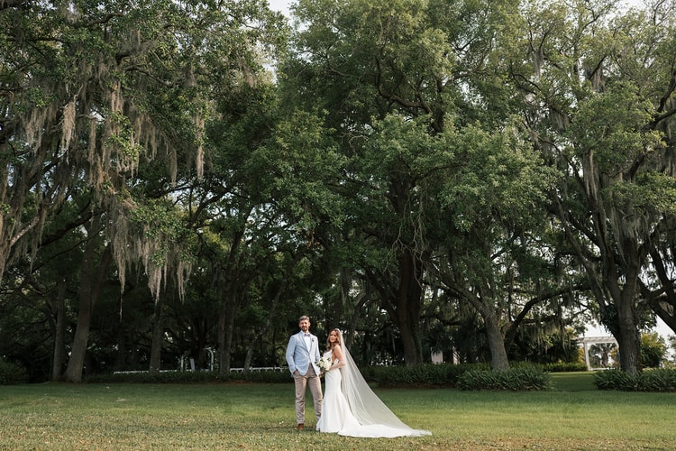 Couple stand together on a wide lawn beneath a canopy of towering moss-draped oaks in soft evening light, wedding photography