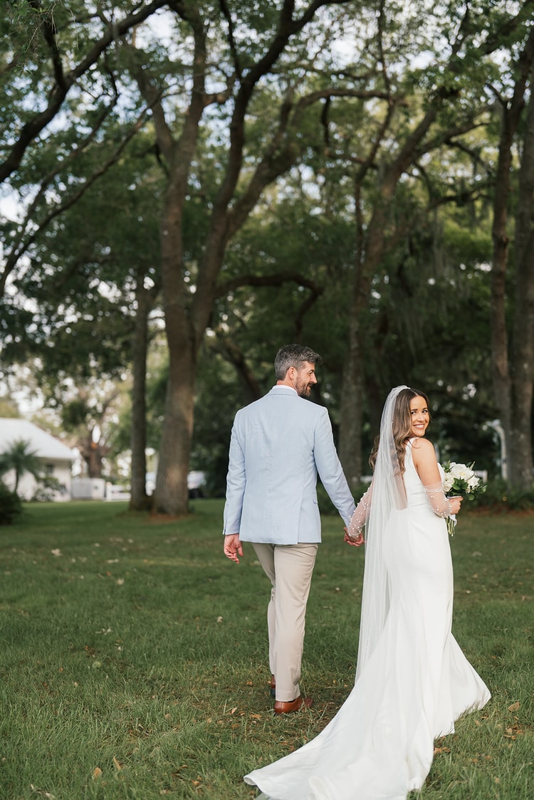 Couple walk hand in hand across a green lawn into a grove of tall trees, bride glancing back and smiling at the groom, wedding photography