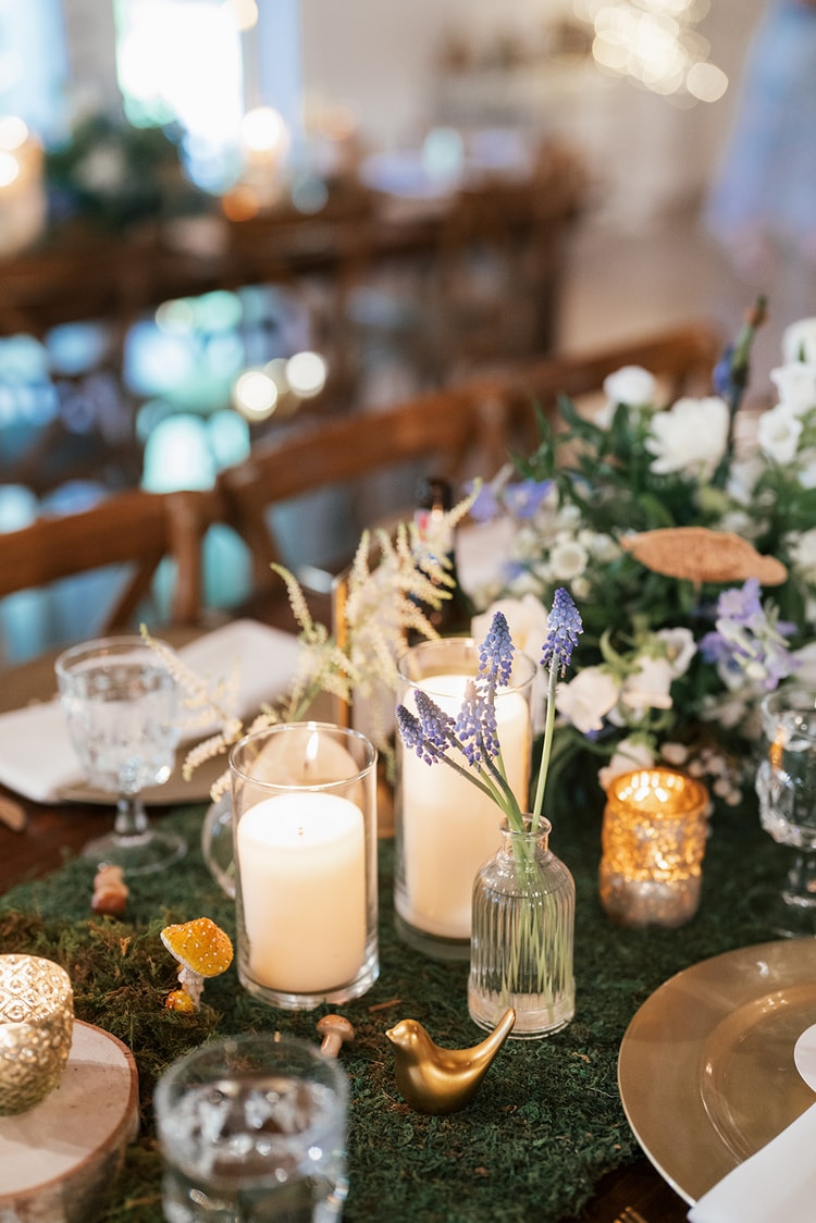 Reception tablescape with moss runner, blue muscari in bud vases, pillar candles, gold toadstool figures, and warm candlelight, wedding details