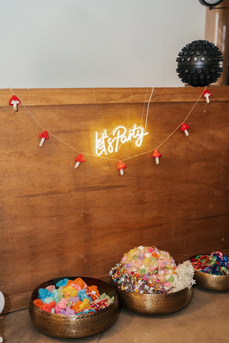 Glowing neon "Let's Party" sign on a wood panel wall above brass bowls filled with colorful party favors and a red mushroom garland, wedding details