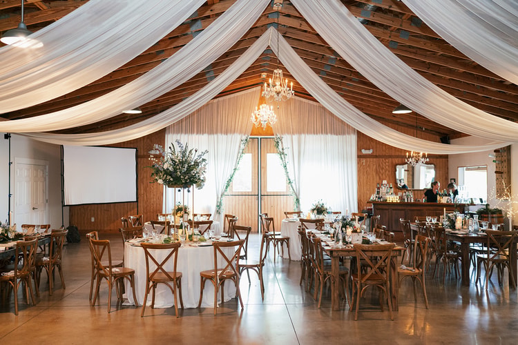 Draped barn reception hall with crystal chandeliers, round and farm tables, cross-back chairs, and tall floral arrangements before guests arrive, wedding details