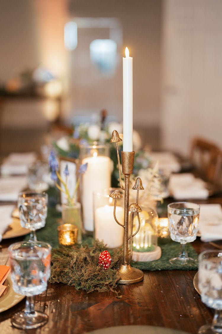 Mushroom-shaped gold candelabra with a lit taper candle on a moss-covered farm table surrounded by pillar candles and blue muscari flowers, wedding details