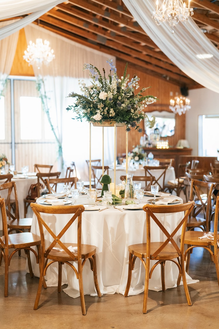 Round reception table with white linen and a tall gold-stand floral centerpiece of white blooms and greenery under draped barn ceilings with chandeliers, wedding details