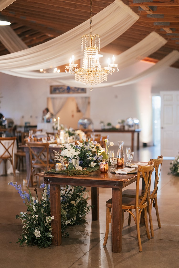 Sweetheart table set beneath a crystal chandelier draped with white fabric, white and blue flowers spilling to the floor in a barn reception space, wedding details