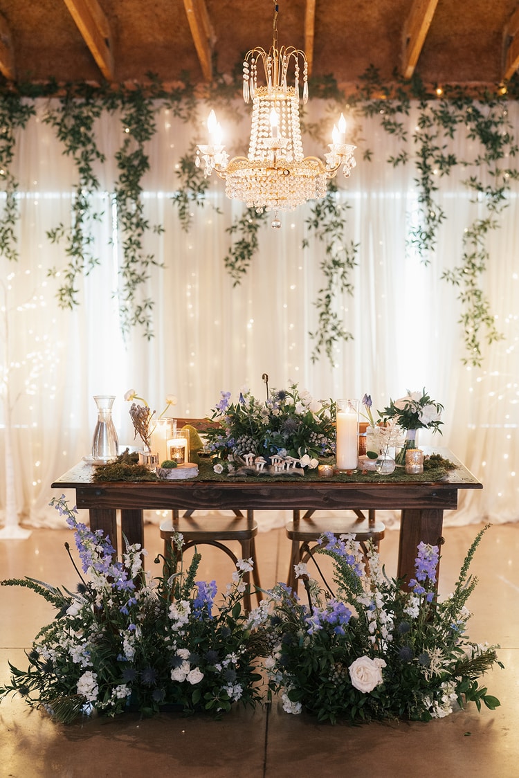Sweetheart table with a lush greenery and blue flower floor arrangement, candles and fairy-light curtain backdrop in a draped barn reception room, wedding details