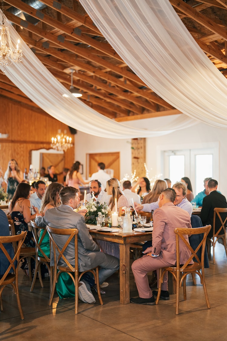 Guests laughing and chatting at farm tables during the reception in a wood-paneled barn with white ceiling draping and chandeliers, wedding photography