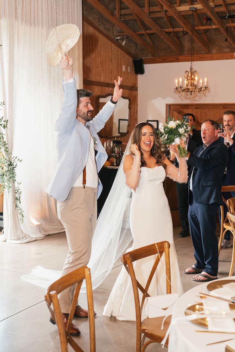 Couple making a jubilant grand entrance into the reception hall, groom raising a hat and bride laughing with bouquet raised, guests cheering around them, wedding photography