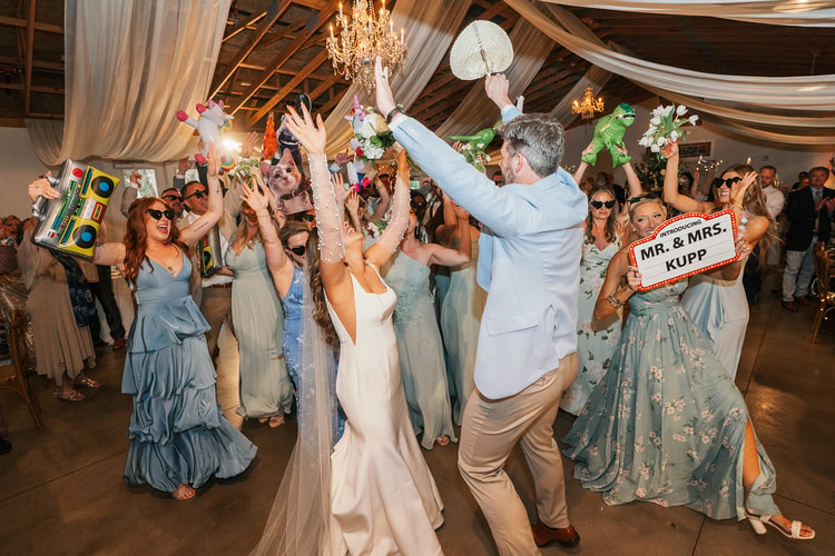 Couple and wedding party erupting in celebration on the dance floor with props raised high, guests in sage dresses and a "Mr. & Mrs. Kupp" marquee sign visible, wedding photography