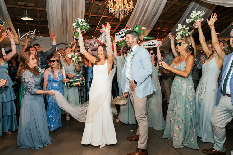 Bride and groom cheering with arms raised while guests in sage dresses hold "Just Married" signs and celebrate around them on the reception dance floor, wedding photography