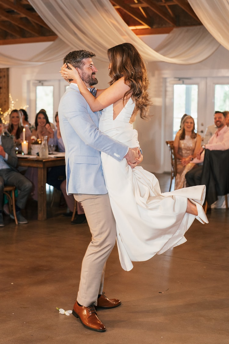 Groom lifting bride off the ground during their first dance, both gazing at each other in a draped barn reception with guests watching from wooden chairs, wedding photography