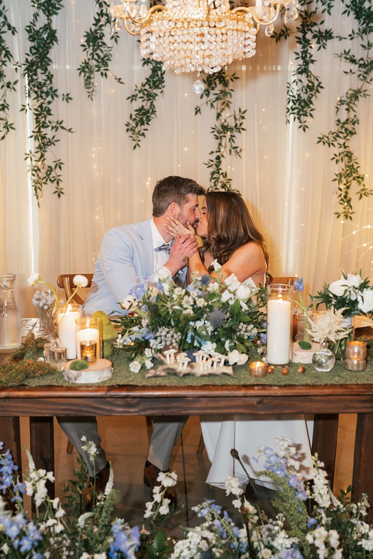 Couple sharing a kiss at the sweetheart table surrounded by blue and white flowers and candles, ivy-draped fairy-light curtain glowing behind them, wedding photography