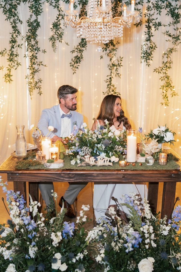 Bride and groom smiling and laughing together at the sweetheart table with blue and white floral arrangements and a glowing fairy-light backdrop, wedding photography