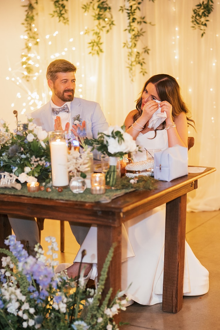 Bride wiping a tear at the sweetheart table while groom applauds beside her, blue and white flowers and candles in the foreground, wedding photography