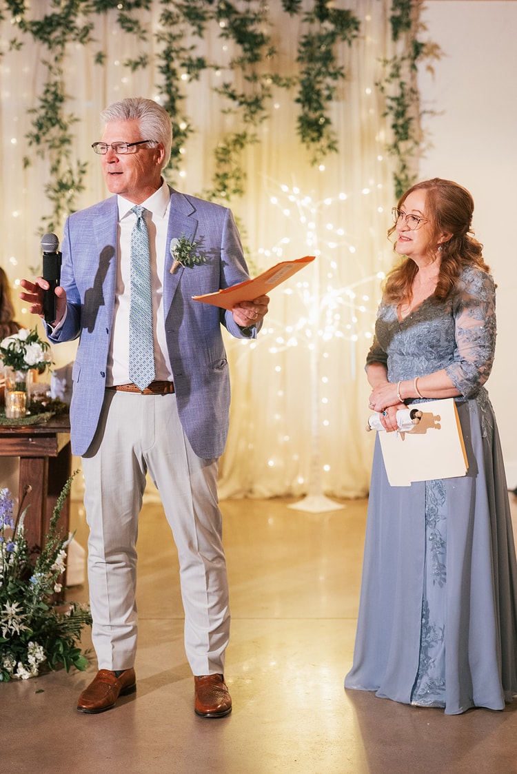 Man in a blue suit delivering a toast with a microphone and notes while a woman in a dusty-blue gown stands beside him before a fairy-light backdrop, wedding photography