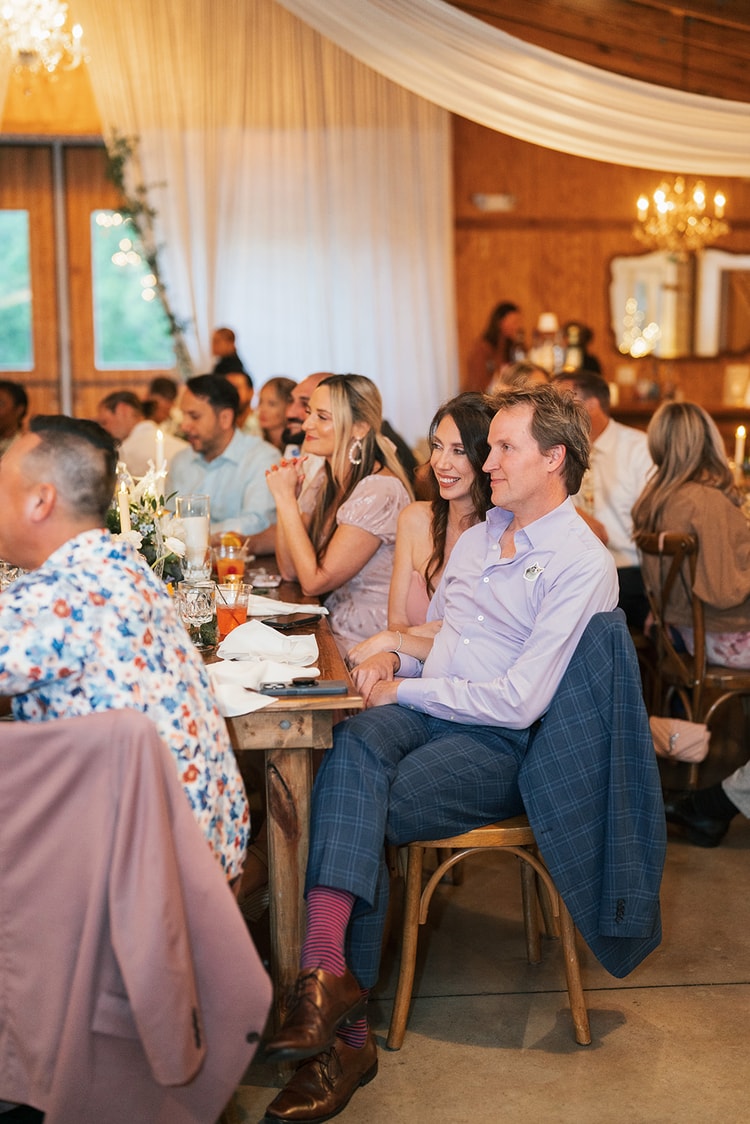 Guests smiling and leaning close at a farm table during the reception in a wood-paneled barn with soft candlelight and chandeliers in the background, wedding photography