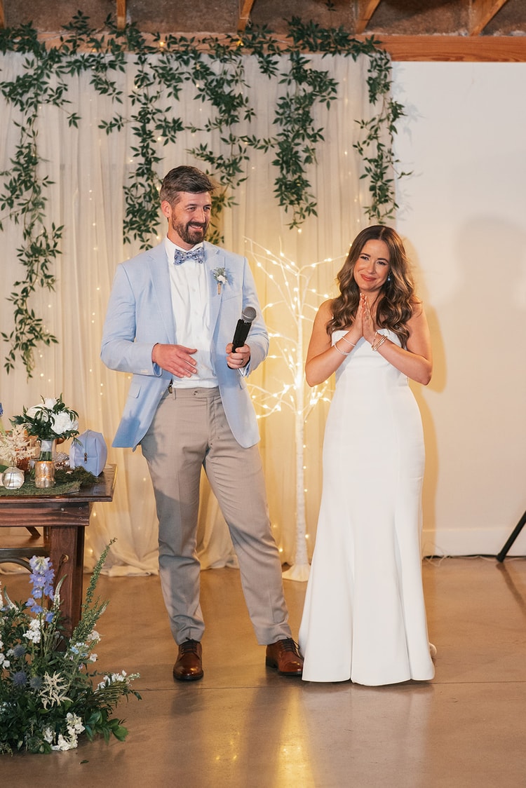 Groom holding a microphone and addressing guests while bride clasps her hands and smiles beside him in front of an ivy and fairy-light curtain backdrop, wedding photography