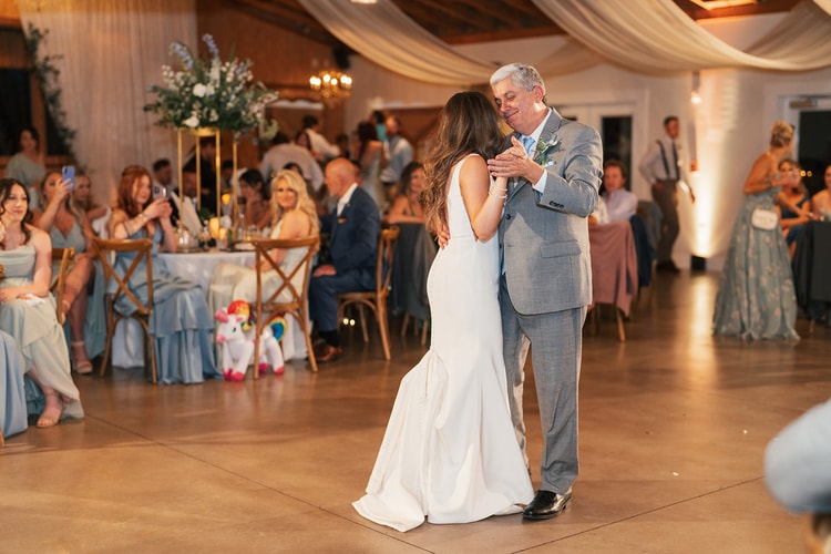 Bride sharing a tender father-daughter dance on the floor of the lit reception barn while guests in sage and blue gowns watch from their seats, wedding photography