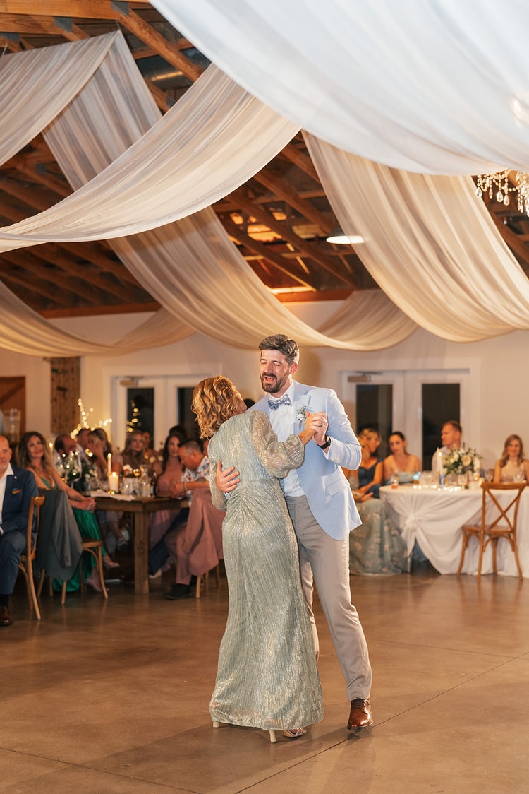 Groom dancing and laughing with his mother in a gold dress on the reception floor under white ceiling draping and chandeliers, wedding photography