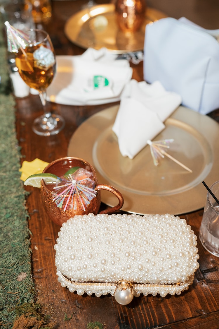 Pearl-covered clutch resting on a moss table runner beside a copper mule mug with a cocktail umbrella and gold charger plate, wedding details