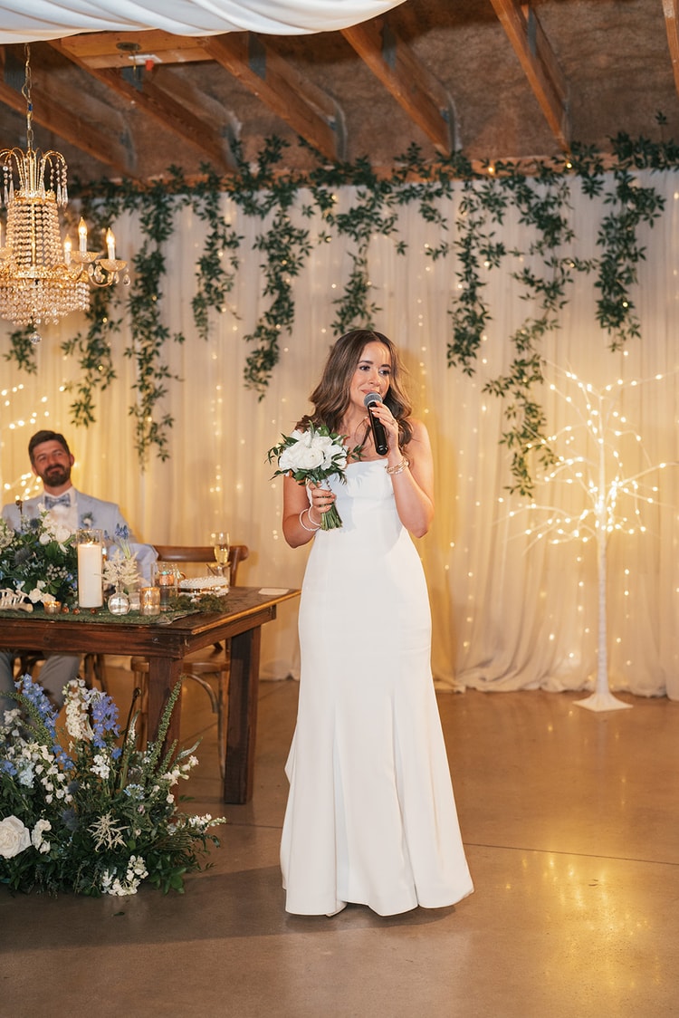 Bride holding a white bouquet and speaking into a microphone at her sweetheart table in front of a glowing ivy and fairy-light curtain backdrop, wedding photography