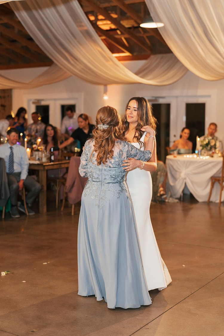 Bride dancing face-to-face with her mother in a dusty-blue embroidered gown on the reception floor while guests look on, wedding photography