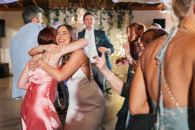 Bride hugging a guest in a pink satin dress on the crowded dance floor while the groom looks on smiling in the background, wedding photography