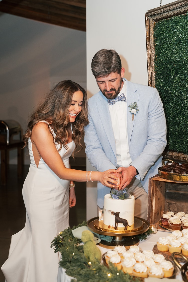 Bride and groom leaning together to cut their white cake decorated with a dog figurine and blue flowers, cupcakes spread around on the dessert table, wedding photography