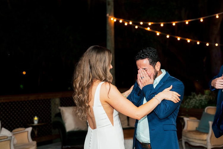 Bride comforting an emotional guest in a navy suit who covers his face with both hands, string lights glowing overhead at an outdoor night reception, wedding photography