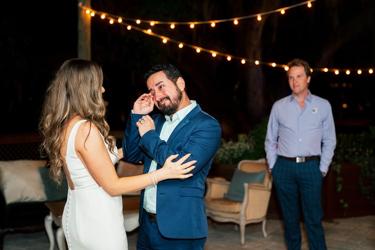 Bride reaching out to wipe a tear from a guest's cheek as he cries at an outdoor night reception under string lights, wedding photography
