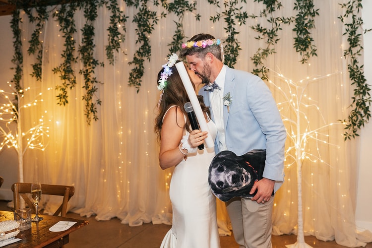 Couple kissing at the sweetheart table, bride wearing a floral crown while groom holds an inflatable dog face prop, fairy-light backdrop behind them, wedding photography