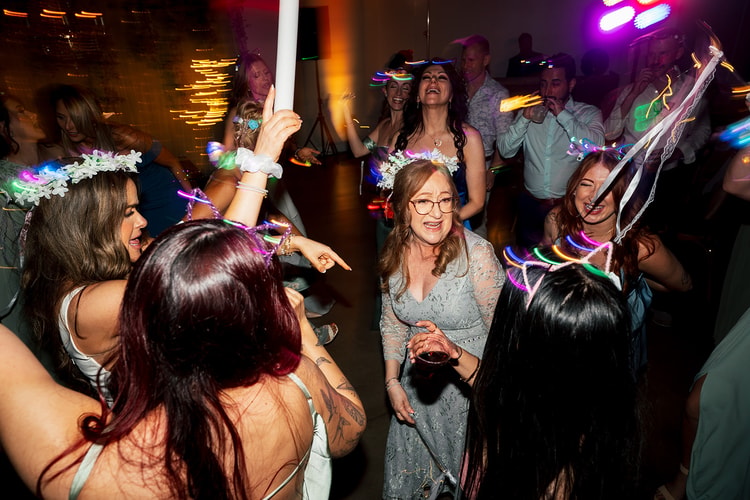 Energetic late-night dance floor packed with guests wearing glow-stick accessories and flower crowns under colorful party lights, wedding photography