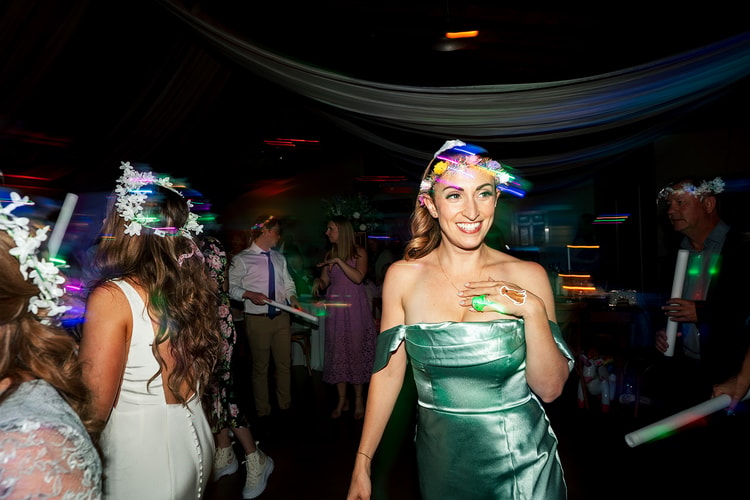 Guest in a green strapless dress dancing and smiling under swirling neon reception lights, flower crown on her head and glow sticks around her, wedding photography