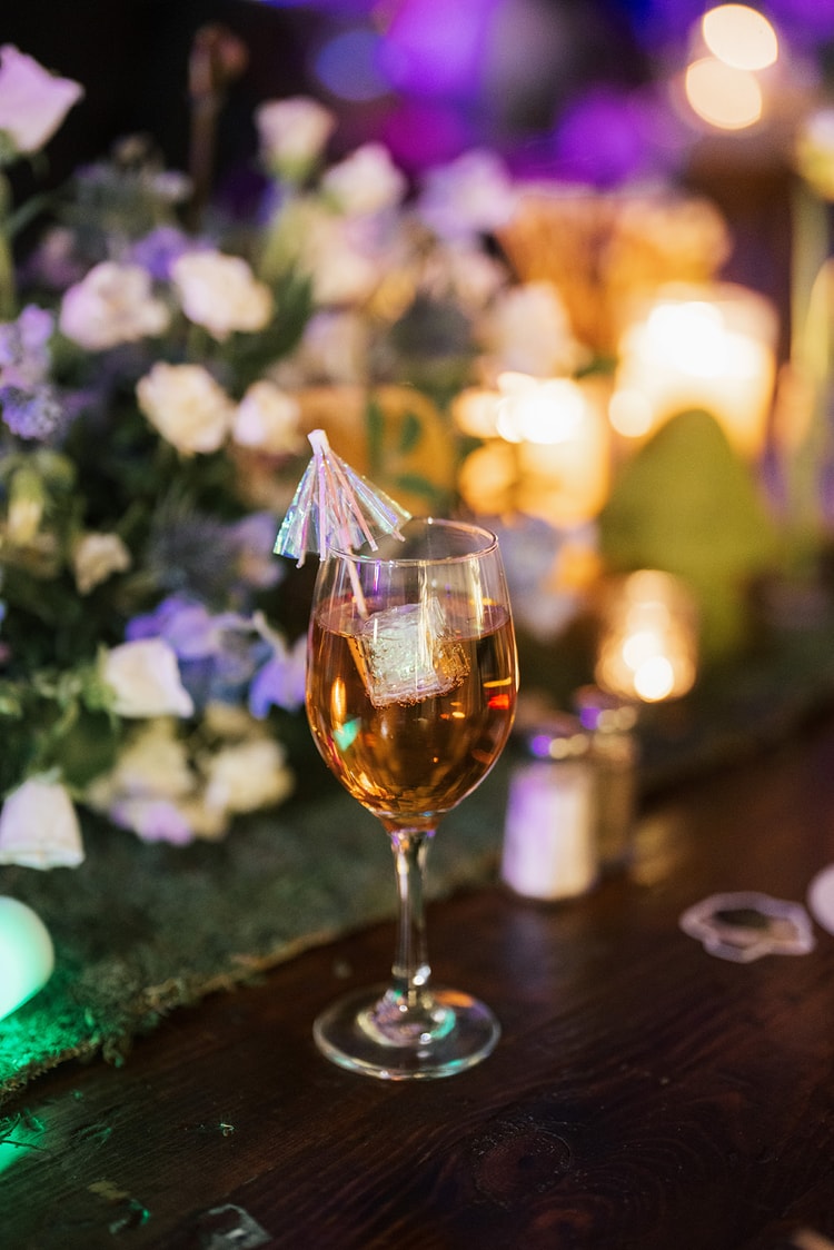 Close-up of a cocktail with an iridescent umbrella straw resting on a moss-covered table beside candles and blue and white floral arrangements, wedding details