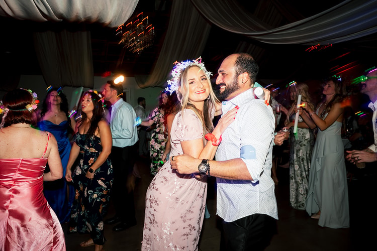 Couple in a floral dress and white shirt dancing close on the reception floor surrounded by guests with glow accessories and flower crowns, wedding photography