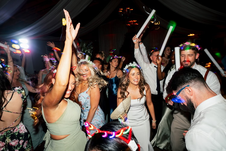 Bride and bridesmaids laughing and dancing wildly with glow sticks and foam wands raised under colorful reception lights, wedding photography