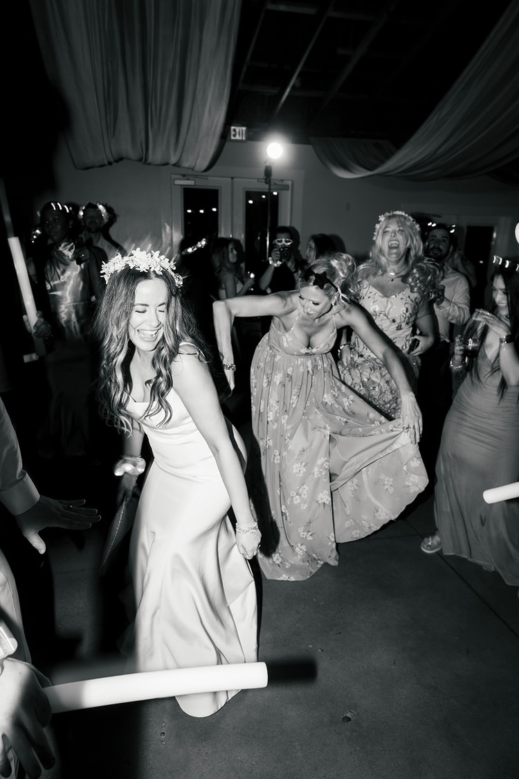 Black-and-white photo of bride in a floral crown bending low during a limbo dance while guests in formal gowns cheer around her, wedding photography