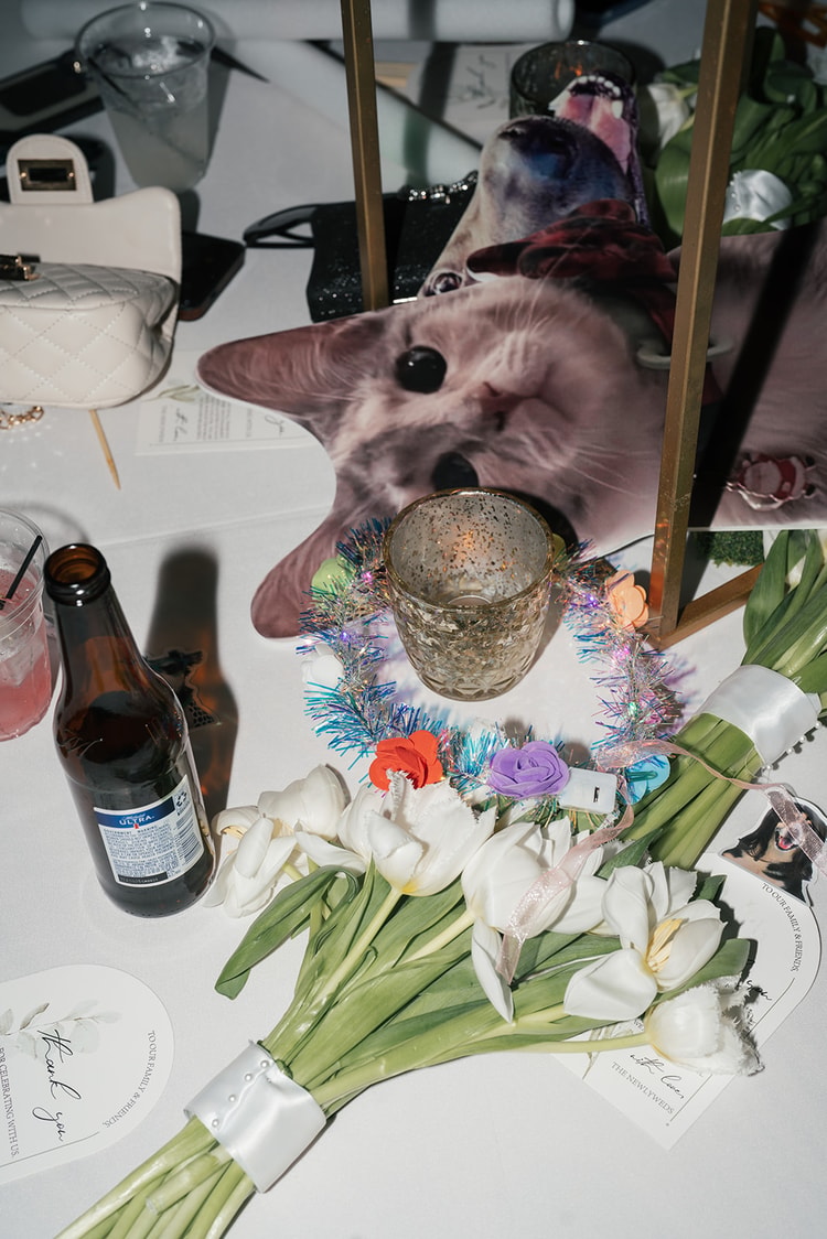 Late-night reception table detail with an oversized cat novelty prop, white tulips, a tinsel garland, and a beer bottle among party debris, wedding details