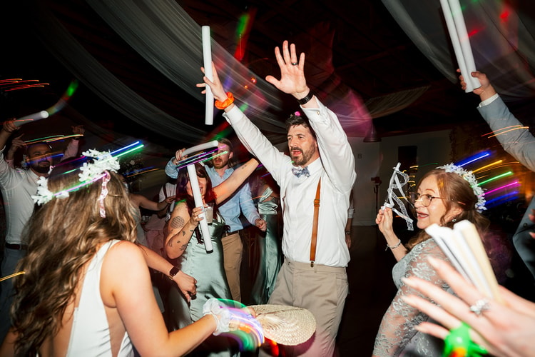 Groom with arms raised dancing at the center of the floor surrounded by guests waving glow sticks and foam wands under spinning reception lights, wedding photography
