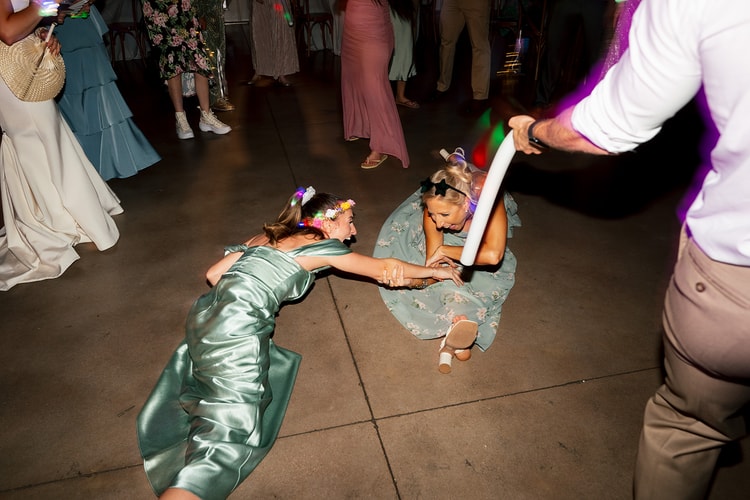 Two bridesmaids in sage dresses sliding on the dance floor holding hands and laughing under colorful reception lighting, wedding photography