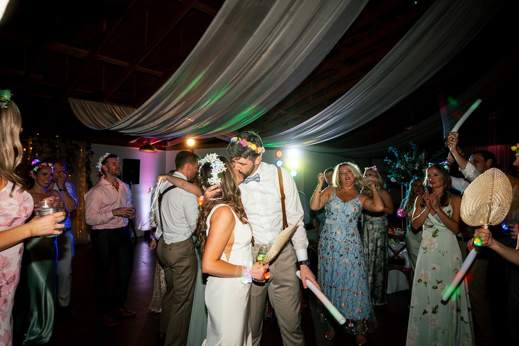 Bride and groom stealing a kiss on the crowded dance floor while guests in sage gowns wave glow sticks and cheer around them, wedding photography