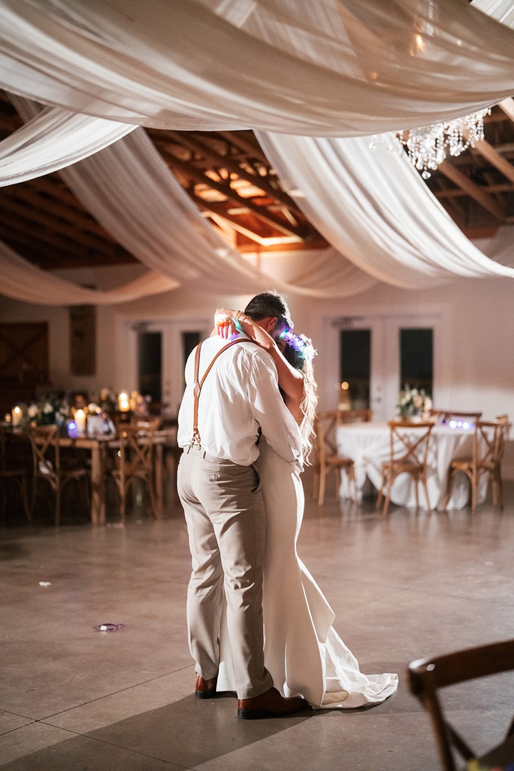 Couple sharing an embrace from behind on the empty dance floor beneath swags of white ceiling draping and a crystal chandelier at the end of the night, wedding photography