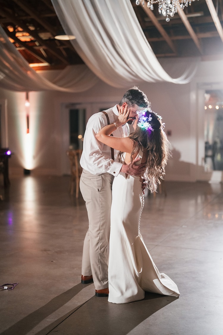 Couple sharing a kiss at the end of the night on the empty dance floor, bride in a lit flower crown, under white ceiling draping and chandeliers, wedding photography