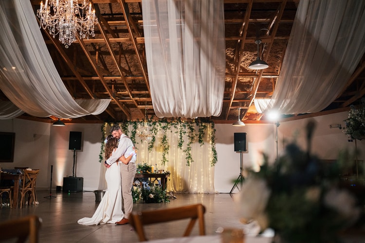 Couple slow-dancing alone on the empty reception floor, sweetheart backdrop glowing with ivy and fairy lights behind them under chandelier and draped ceiling, wedding photography