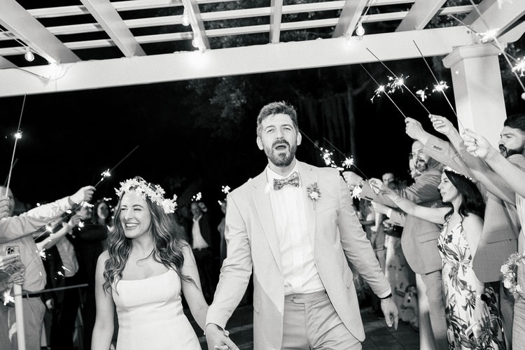 Black-and-white sparkler exit under a pergola, bride in a floral crown and groom in suspenders beaming as guests hold sparklers on both sides, wedding photography