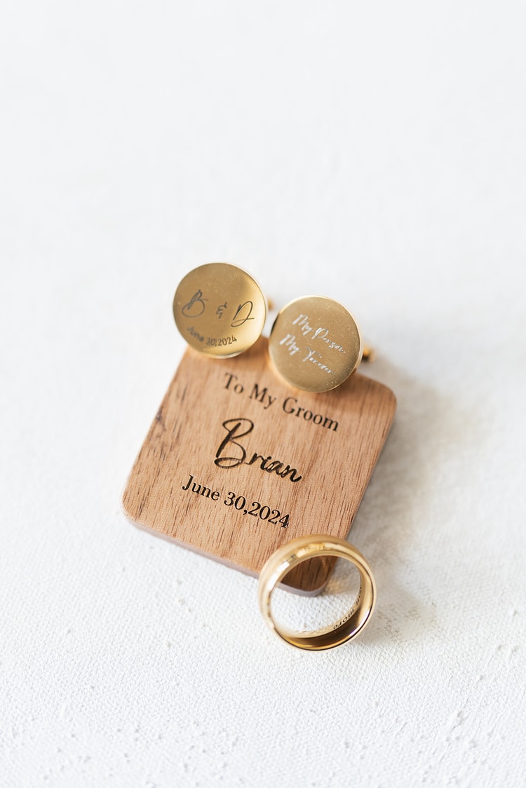 Gold cufflinks engraved with initials and a date resting on a wooden gift box beside a gold wedding band