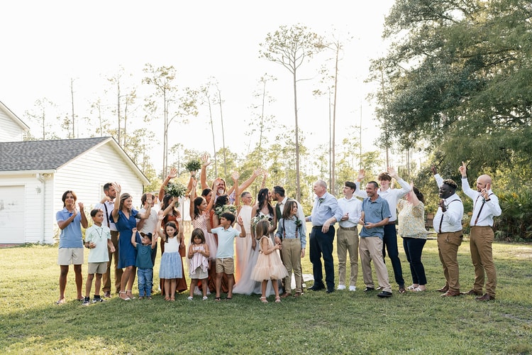 Guests cheer and toss arms in the air around the couple on the lawn at sunset