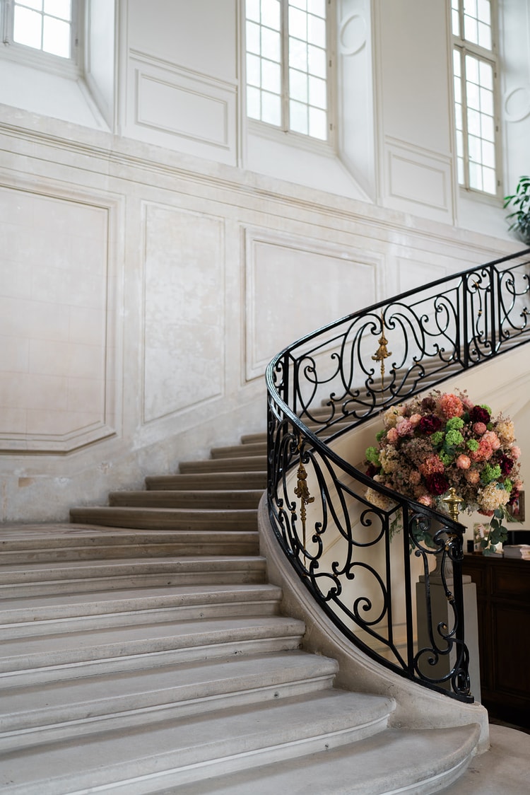Curved stone staircase with scrolled wrought-iron railing and a lush flower arrangement inside Domaine du Château d'Ermenonville
