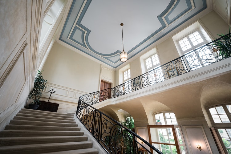 Grand staircase hall of Domaine du Château d'Ermenonville, stone steps rising past an iron balustrade to a bright gallery under a blue-panelled ceiling