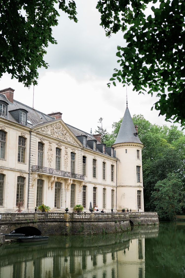 Turret and sculpted facade of Domaine du Château d'Ermenonville reflected in the still green moat, framed by dark summer foliage.